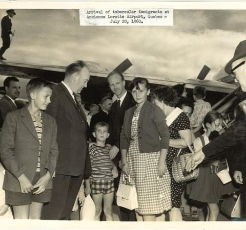 A sepia-toned photo of a large group of formally-dressed, light-skinned people on a tarmac with a plane behind them.