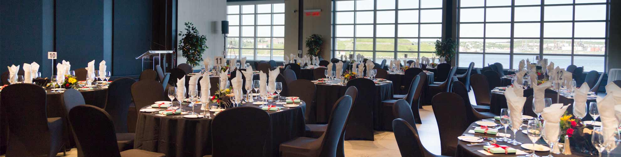 Une salle dont les fenêtres allant du sol au plafond offrent une vue sur le port et l’île et des tables de banquet ovales avec des chaises et des ustensiles de cuisine.