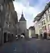 L'image montre une scène de rue animée dans une ville européenne avec une architecture historique et une ligne de tramway. Les gens marchent sous les vieux bâtiments et les arcades, ce qui crée une atmosphère de vie quotidienne et d'histoire.