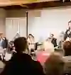 L'image montre une table ronde dans une salle de réunion animée. Une oratrice se tient devant le public qui écoute attentivement. L'ambiance est sérieuse et engagée, reflétée par le public intéressé et les participants au panel concentrés.