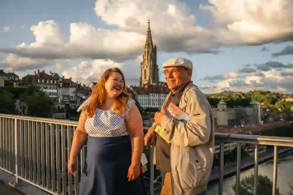 Deux personnes partagent un rire chaleureux sur un pont, dans un décor pittoresque de vieille ville avec une flèche d'église en arrière-plan. La lumière chaude du soleil couchant renforce l'atmosphère joyeuse et légère du moment.