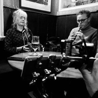 A woman playing concertina and a man playing whistle around a pub table with a guitar headstock in the foreground.