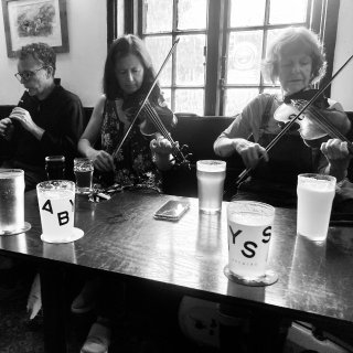 A man playing tin whistle and two women playing fiddle at a pub table.