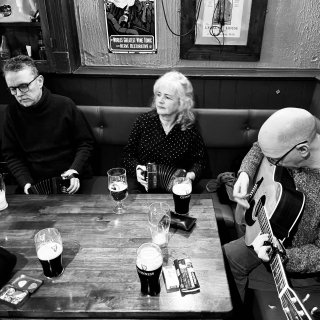 A man playing concertina, a woman playing concertina, and a man playing guitar at a table in a pub corner.