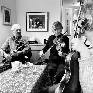 A piper and a fiddler playing at a kitchen table while another fiddler listens.