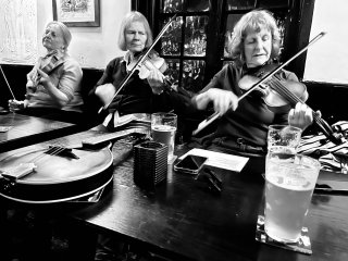 Three women in a row playing fiddle in front of a table with a mandolin on it.