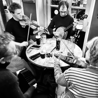 Four musicians playing around a circular table; two fiddles, banjo and concertina.