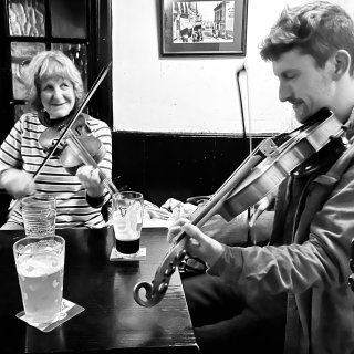 A woman playing fiddle and a man playing fiddle at a table in the corner of a pub.