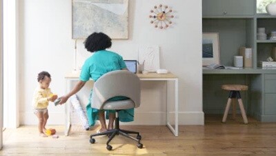 A mother hands a toy to her toddler while sitting at her desk with laptop. 