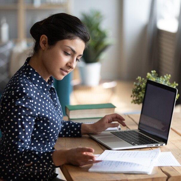 A woman wearing a polka-dotted blouse studies at her desk with her laptop and notes in front of her.