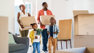 A photo of a family standing in the living room of a house. They are holding moving boxes and there are moving boxes placed in the living room.