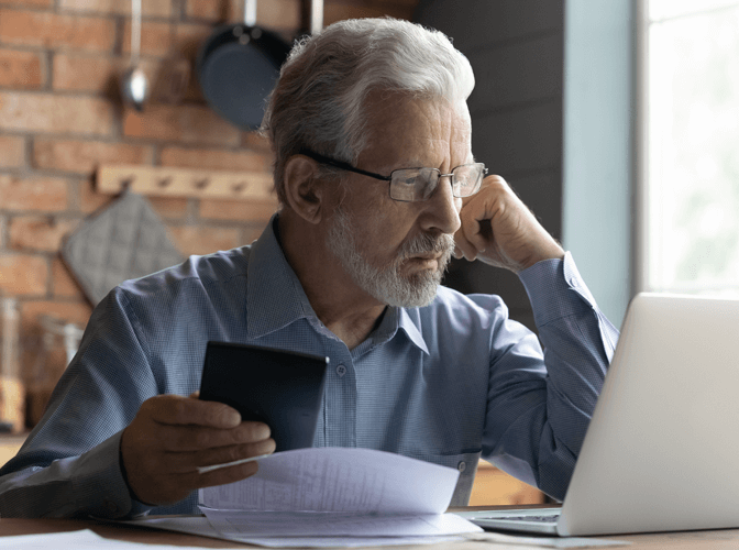 Man considers something on a laptop with concern while holding a calculator and what might be a financial statement.