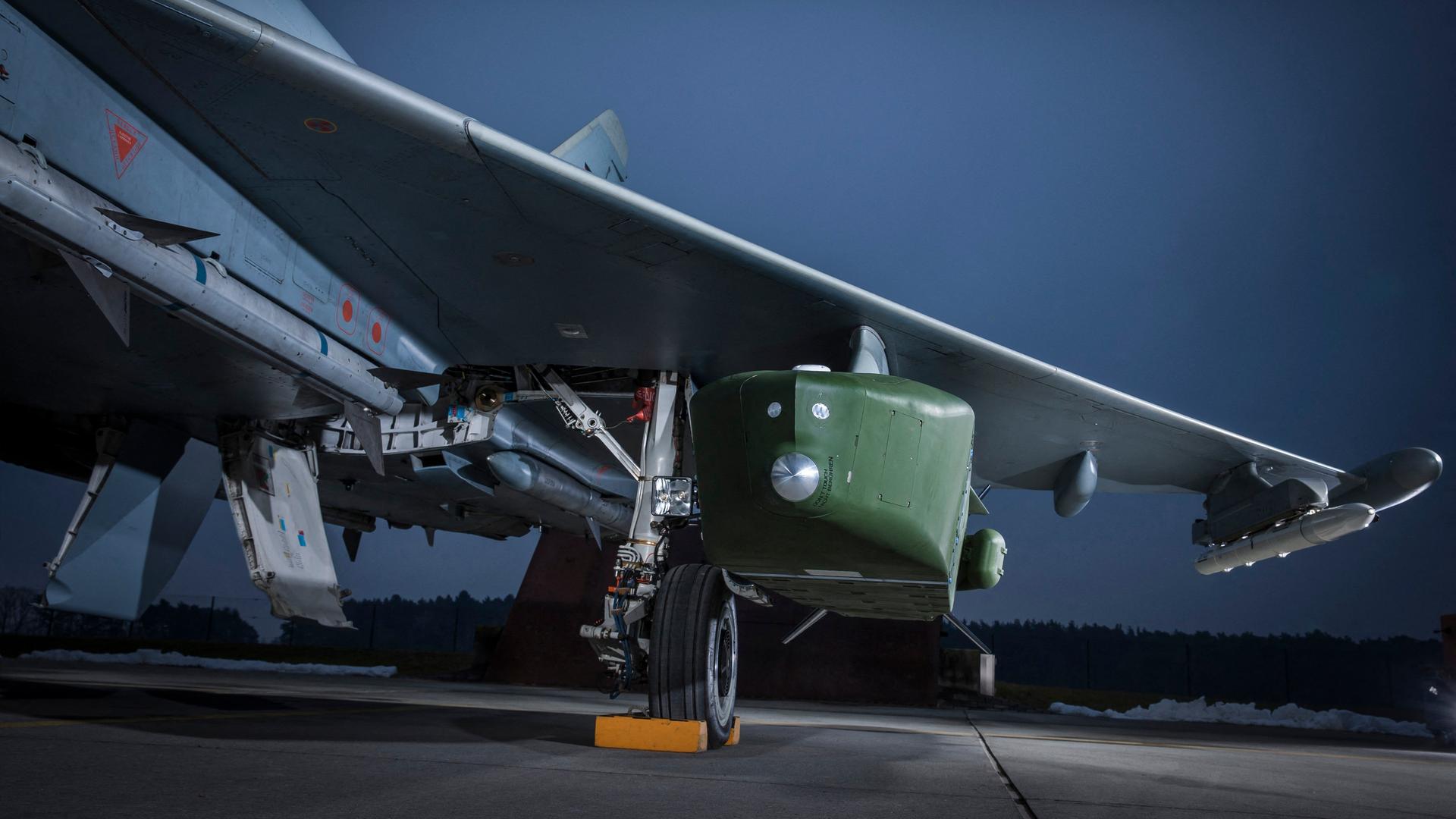 Der Marschflugkörper Taurus KEPD 350 unter einem Bundeswehr-Kampfjet. Der Himmel im Hintergrund leuchtet bläulich. Es ist Abend. 