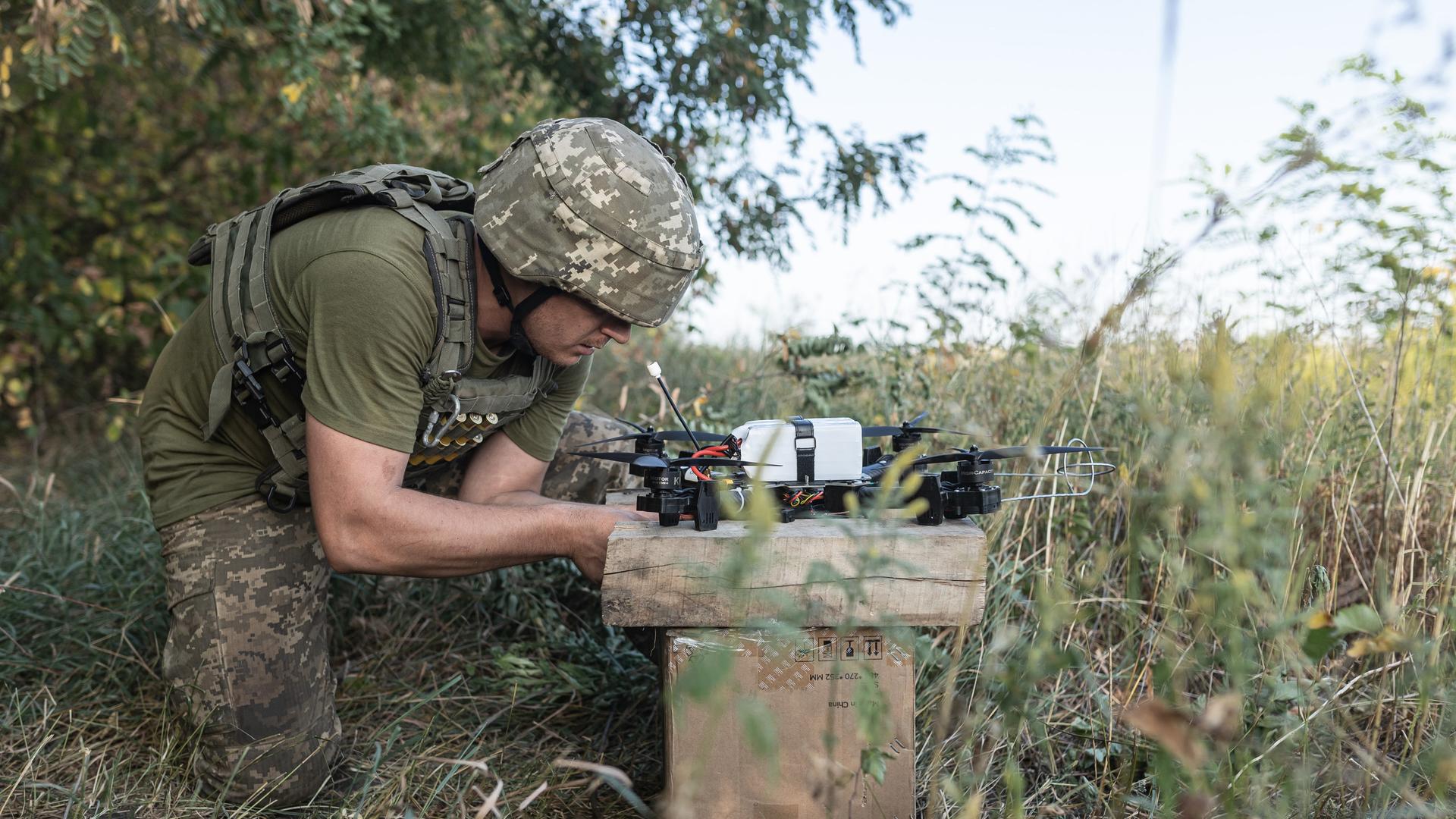 Ein ukrainischer Soldat bückt sich zu einer Drohne, die mit etwas beladen ist. Drumherum sind Wald und Büsche.