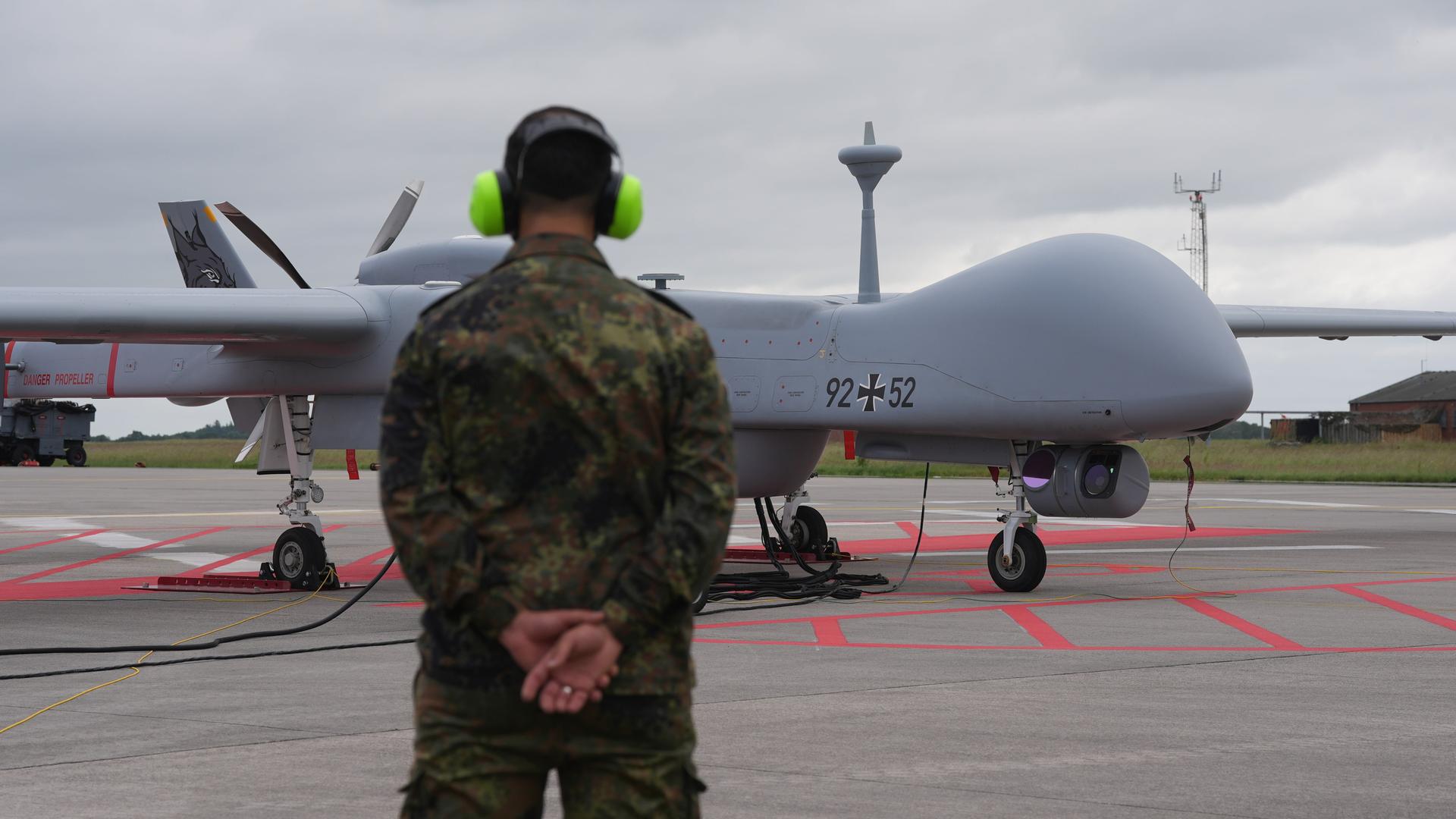 Eine Heron-Drohne der Bundeswehr steht auf einem Flugfeld. Davor ein Soldat mit Gehörschutz.