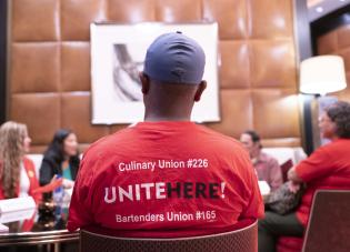 Vários trabalhadores sentam e conversam com a secretária interina Su no The Venetian Resort. A camisa vermelha de um trabalhador, vista de trás, tem os dizeres “Culinary Union #226. Inscreva-se aqui! Sindicato dos Bartenders #165”. 