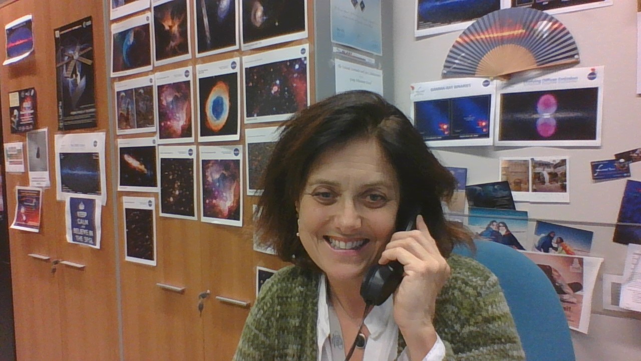 A woman with short brown hair talks on the phone in an office with images from IXPE behind her.