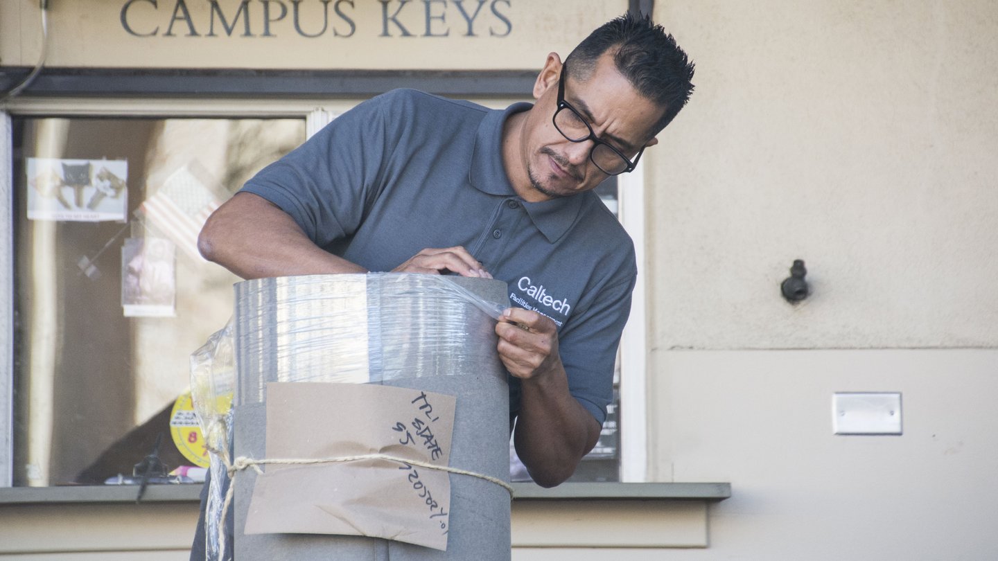 Carpenter Supervisor Chay tearing plastic packaging materials from gray package. He is standing in front of window with sign above reading "Campus Keys"