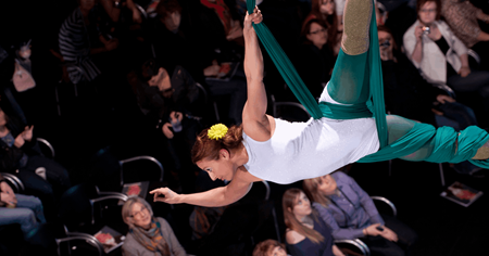 A circus artist wrapped in aerial silks flies over a seated audience.