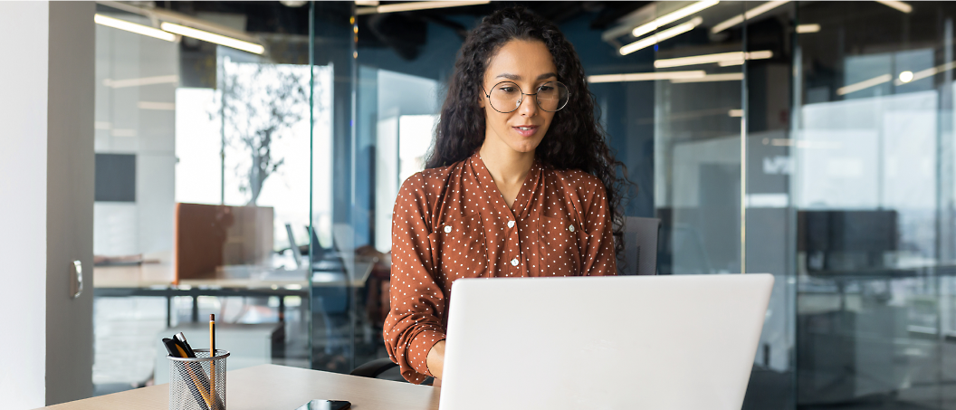 A woman with curly hair, wearing glasses and a polka dot shirt, works on her laptop in a modern office.