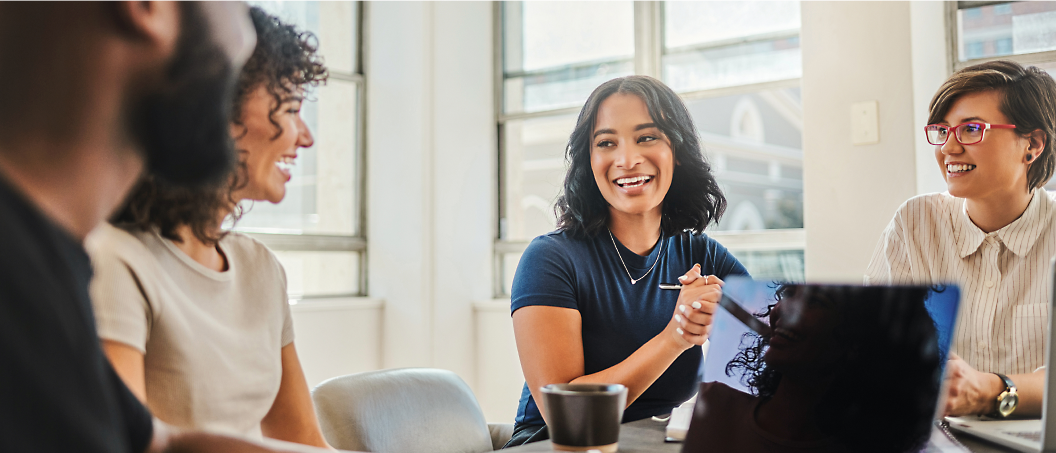 A diverse group of colleagues engaging in a lively discussion around a table in a sunny, modern office setting.