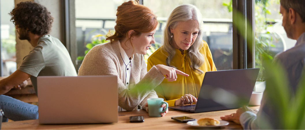 Two women, one younger and one older, are looking at a laptop screen together in a cafe setting during daylight.