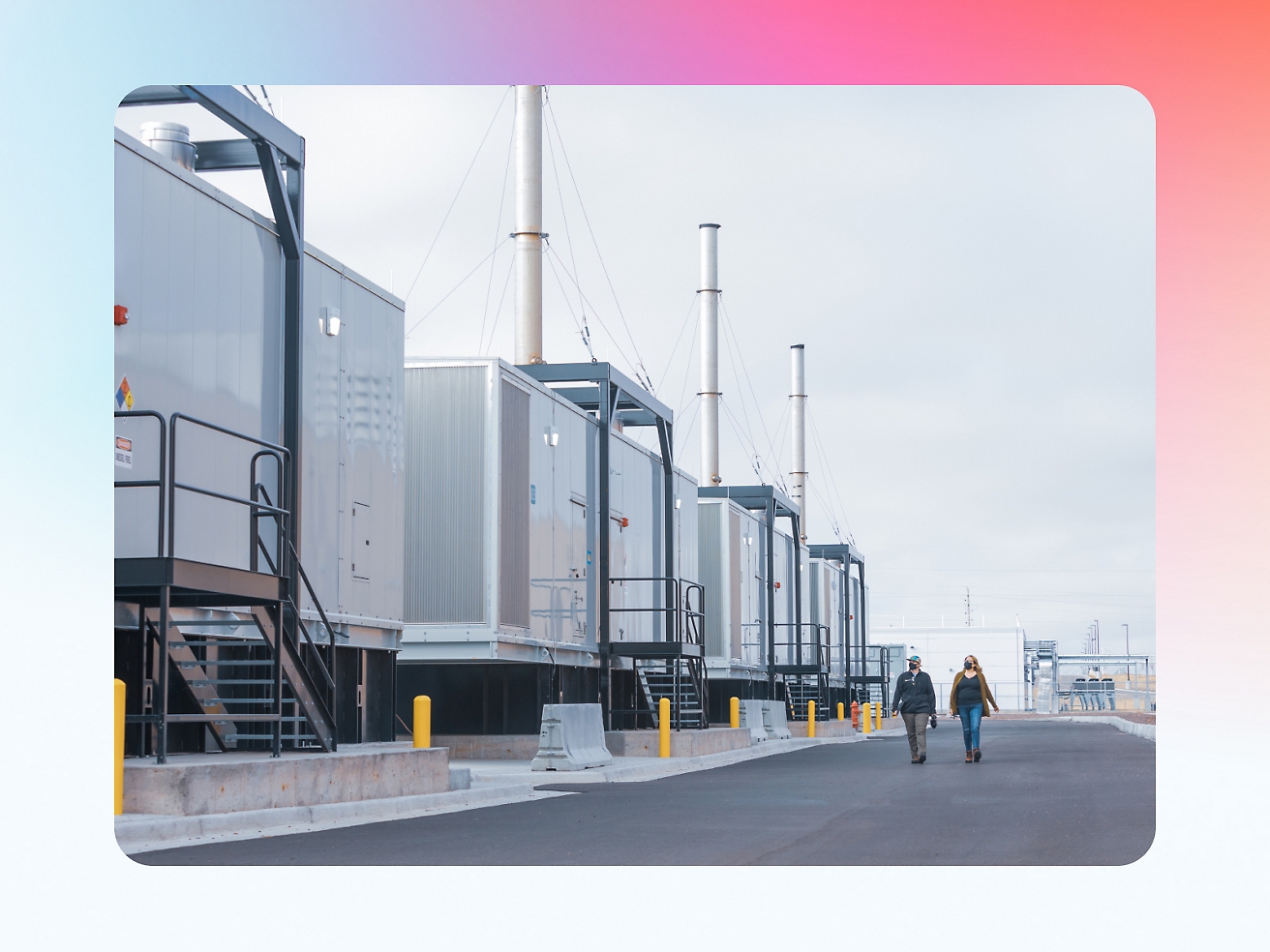 Two people walking by a row of industrial containers with tall chimneys at a facility, under an overcast sky.