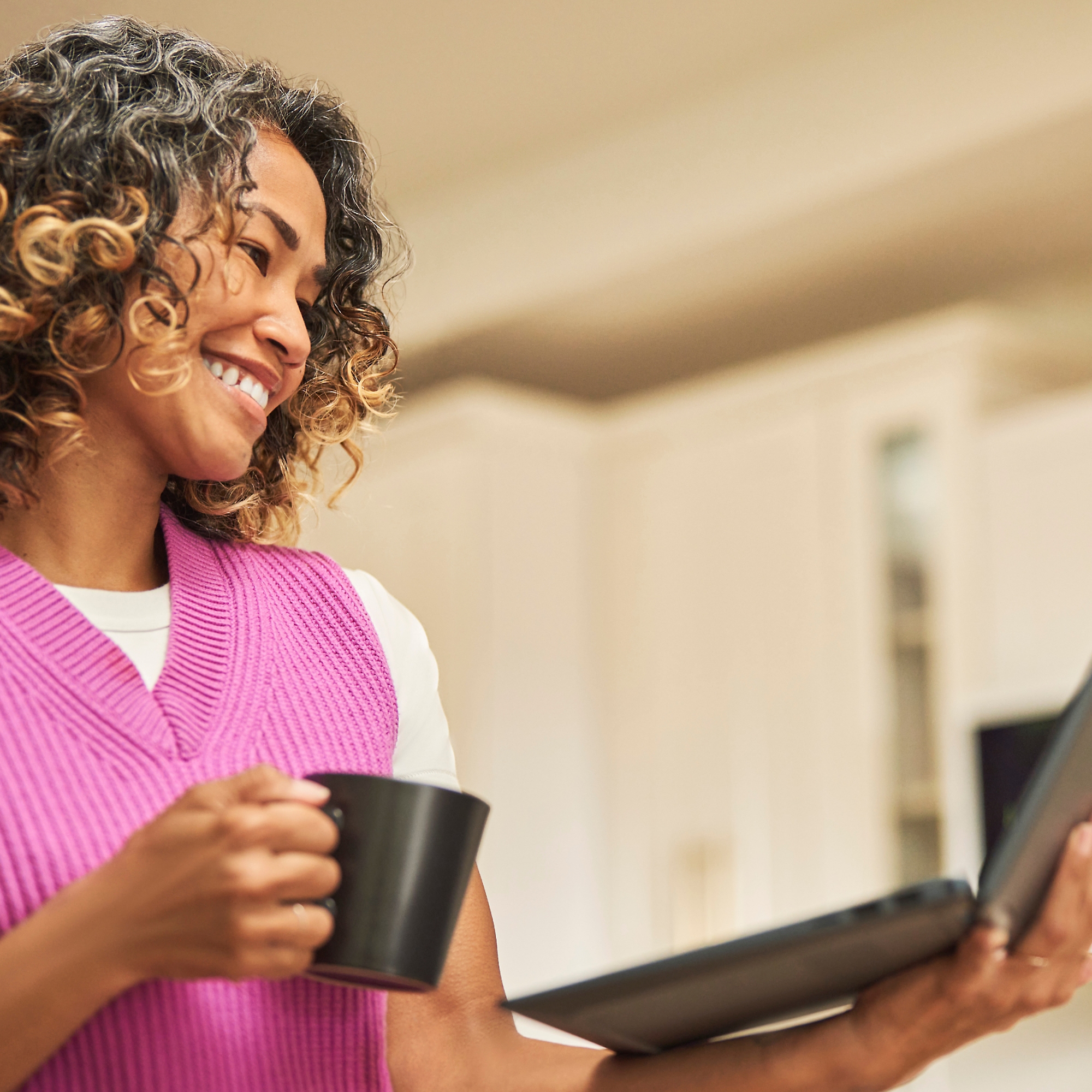 Moteris, besišypsanti su kavos puodeliu vienoje rankoje ir nešiojamuoju kompiuteriu kitoje