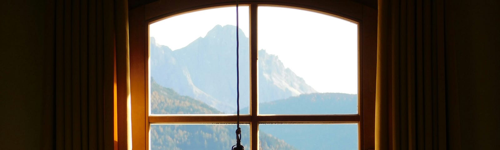 A view through a small window, divided into four by thin strips of wood, showing distant hazy blue hills and a mountain peak. Pink flowers in foreground outside the window. The walls around the window are dark, emphasizing the light outside.