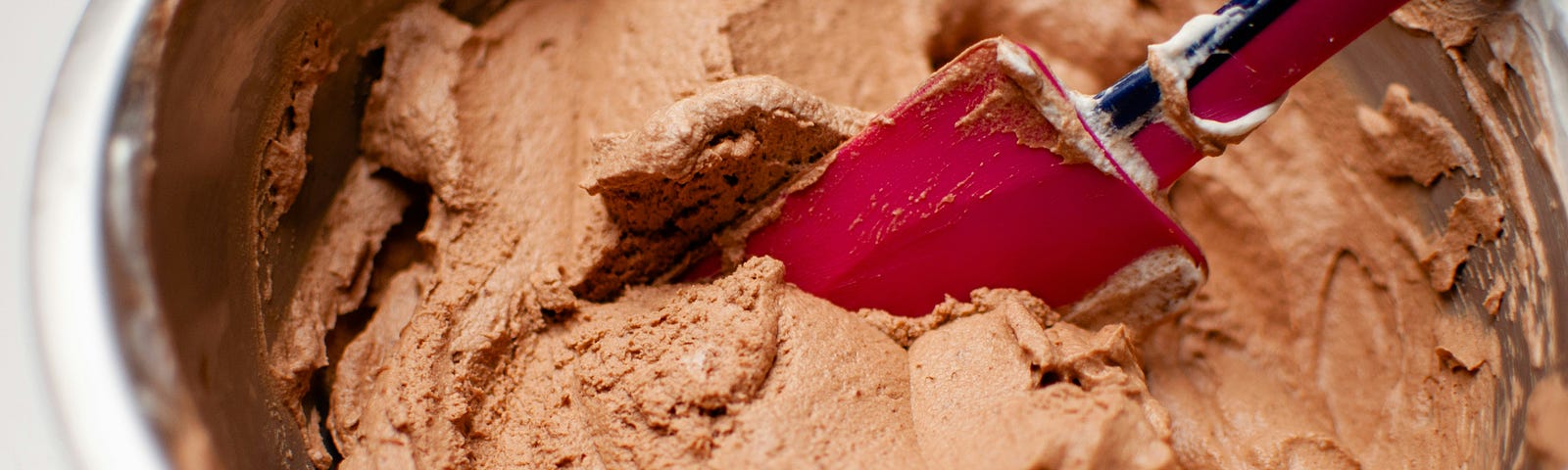 A red spatula is resting in the chocolate batter in a metal baking bowl