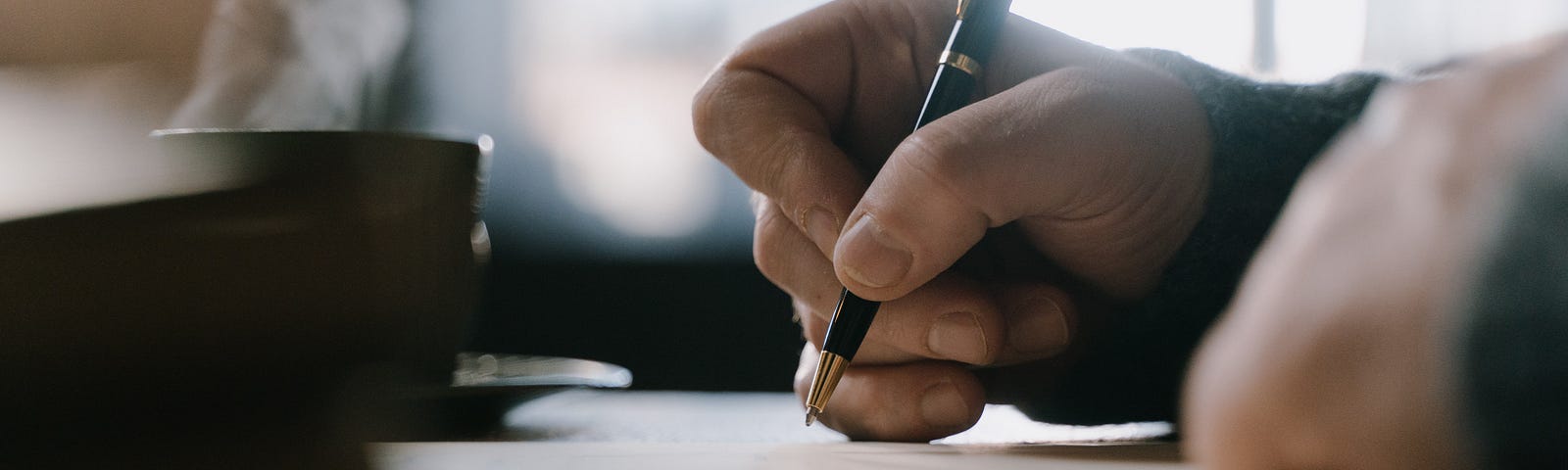 A side POV of hands holding a pen about to write on paper. Further back is a coffee cup with steam coming out of it.