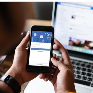 A man holds his cell phone in front of him, the Facebook login screen is displayed. In the background, his Facebook news feed is pulled up on his laptop.