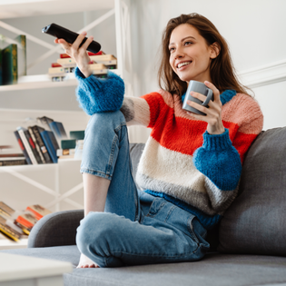 A woman sits on her couch, holding a mug and the remote.