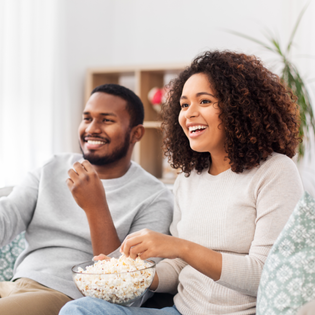 A man and woman sit next to each other on the couch, eating popcorn.