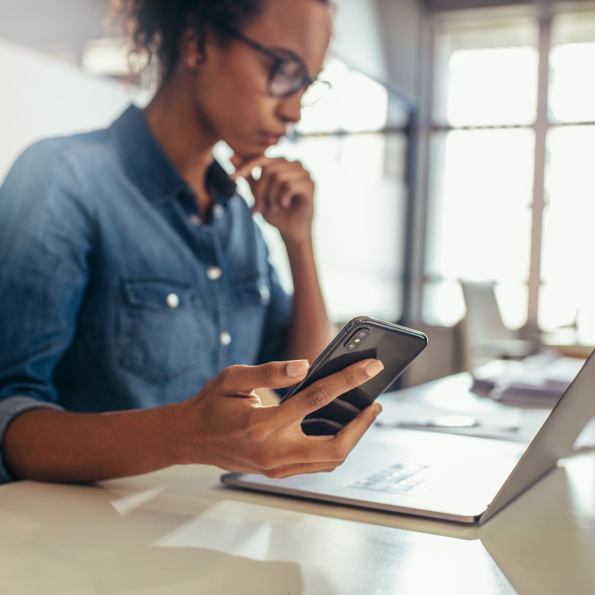 A young Black woman holds her smartphone in her right hand while looking at her laptop screen