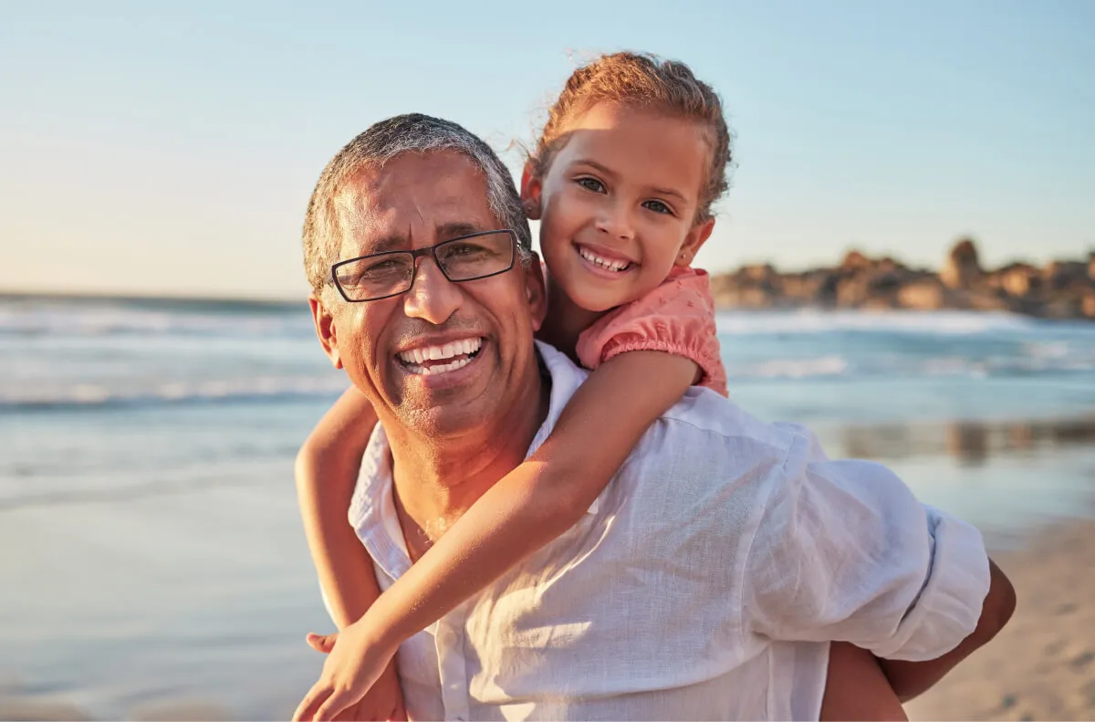 A man in glasses carries a child on his back at the beach