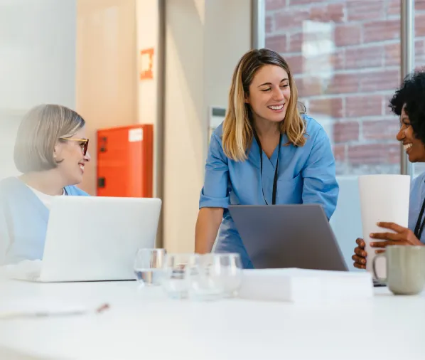 Three women in blue uniforms, smiling and working together with laptops at a table.