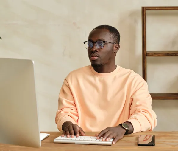 A man in glasses and a light peach sweater, working on a computer at a wooden desk.