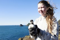 A young person holds a juvenile puffin while standing atop a sea cliff.