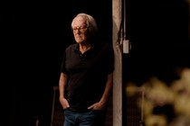 A man wearing a black shirt and jeans leans against a wood post.