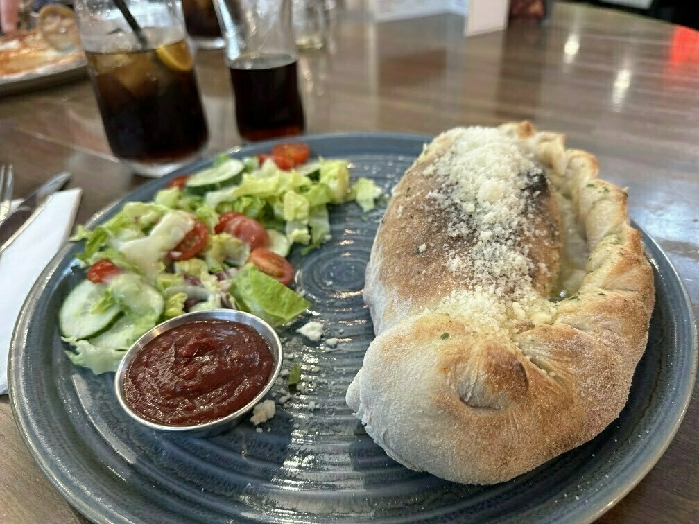 A calzone topped with grated cheese is served on a plate with a fresh salad and a side of tomato sauce, accompanied by two glasses of cola.