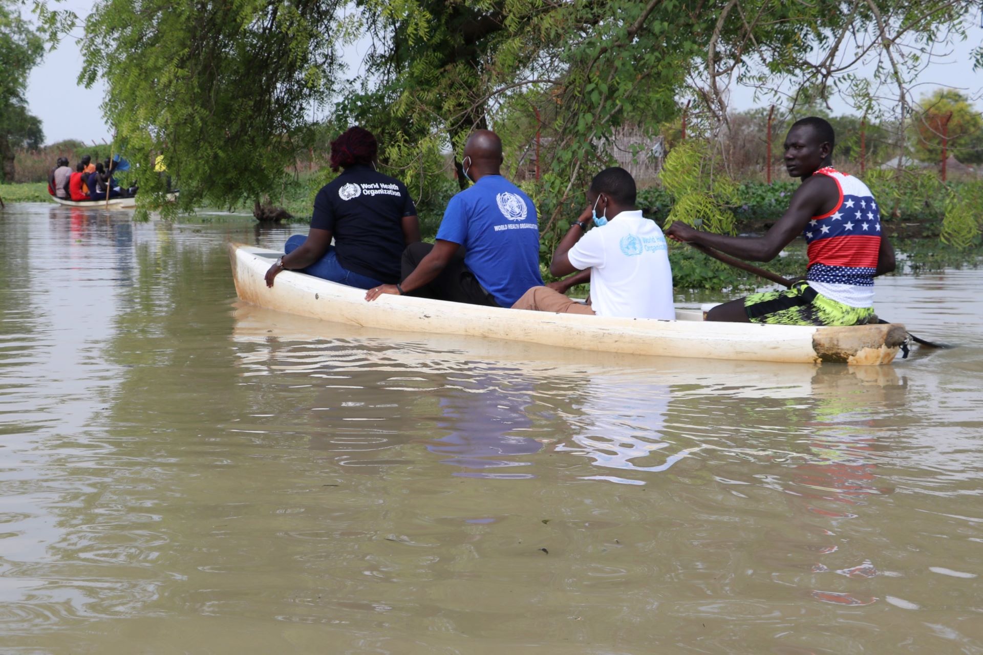 Four people in a canoe, three of whom are wearing WHO T-shirts, travelling on flooded ground.
