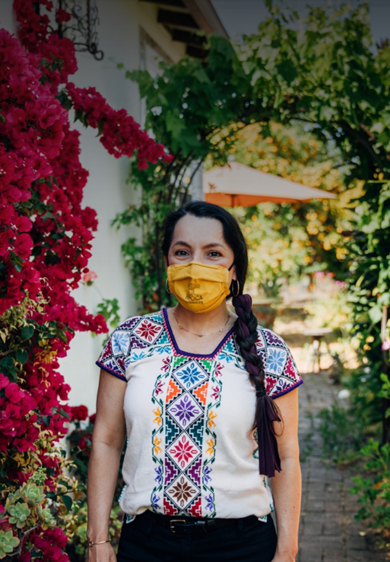 A woman in a colorful blouse, wears a yellow face mask bearing the NFOCA logo in her garden.