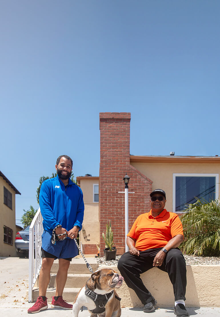 A man in a blue sweatshirt hold his bulldog on a leash and stands next to a man in an orange T-shirt and hat.