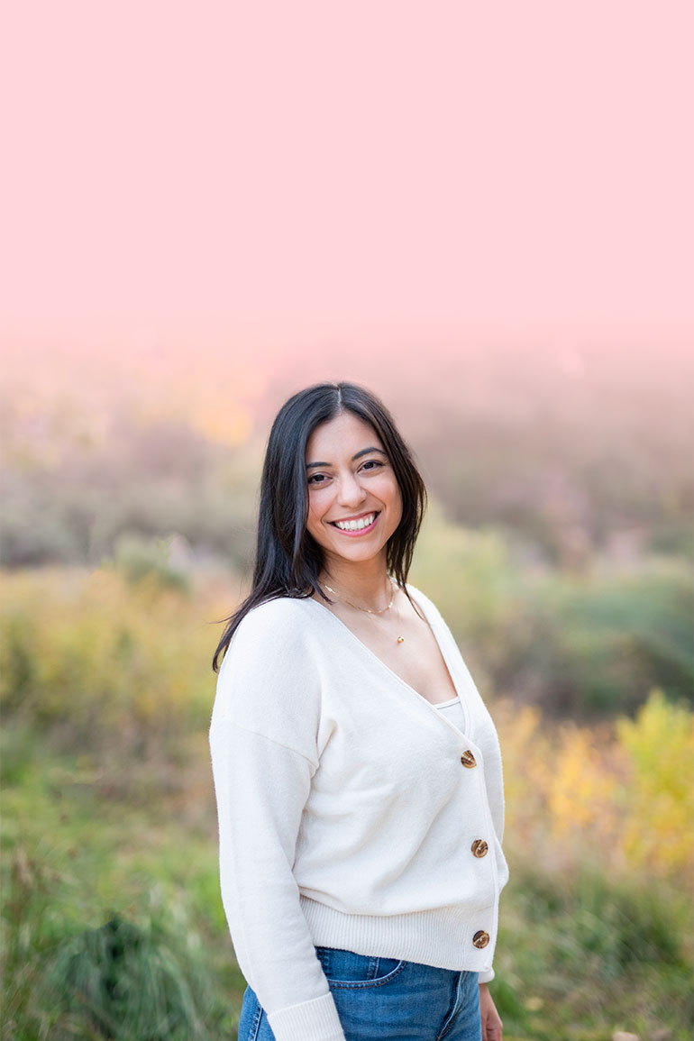 A woman stands outside and smiles, wearing a white blouse.