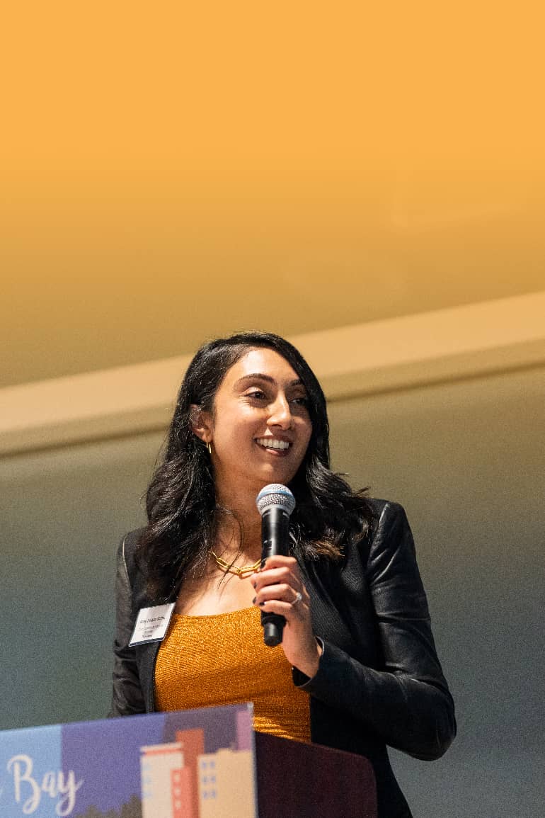 Ruby Bolaria Shifrin holds a microphone while speaking at an event.