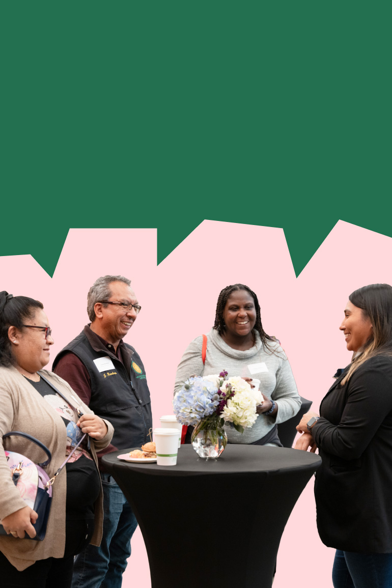 Four people laugh as they stand around a cocktail table. Others chat behind them, amidst a vibrant backdrop of art work.