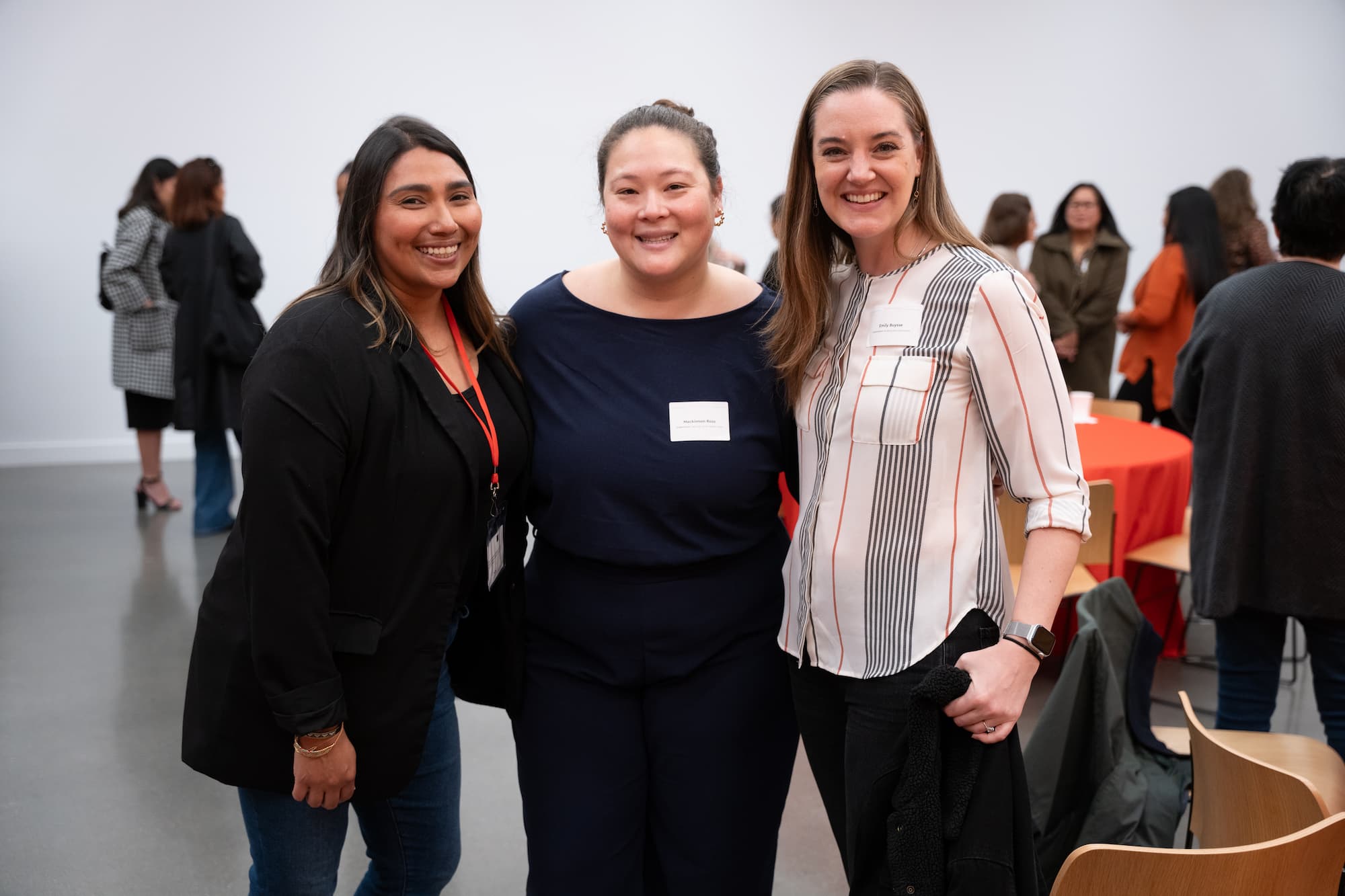 Three women pose together with tables and people in the background.