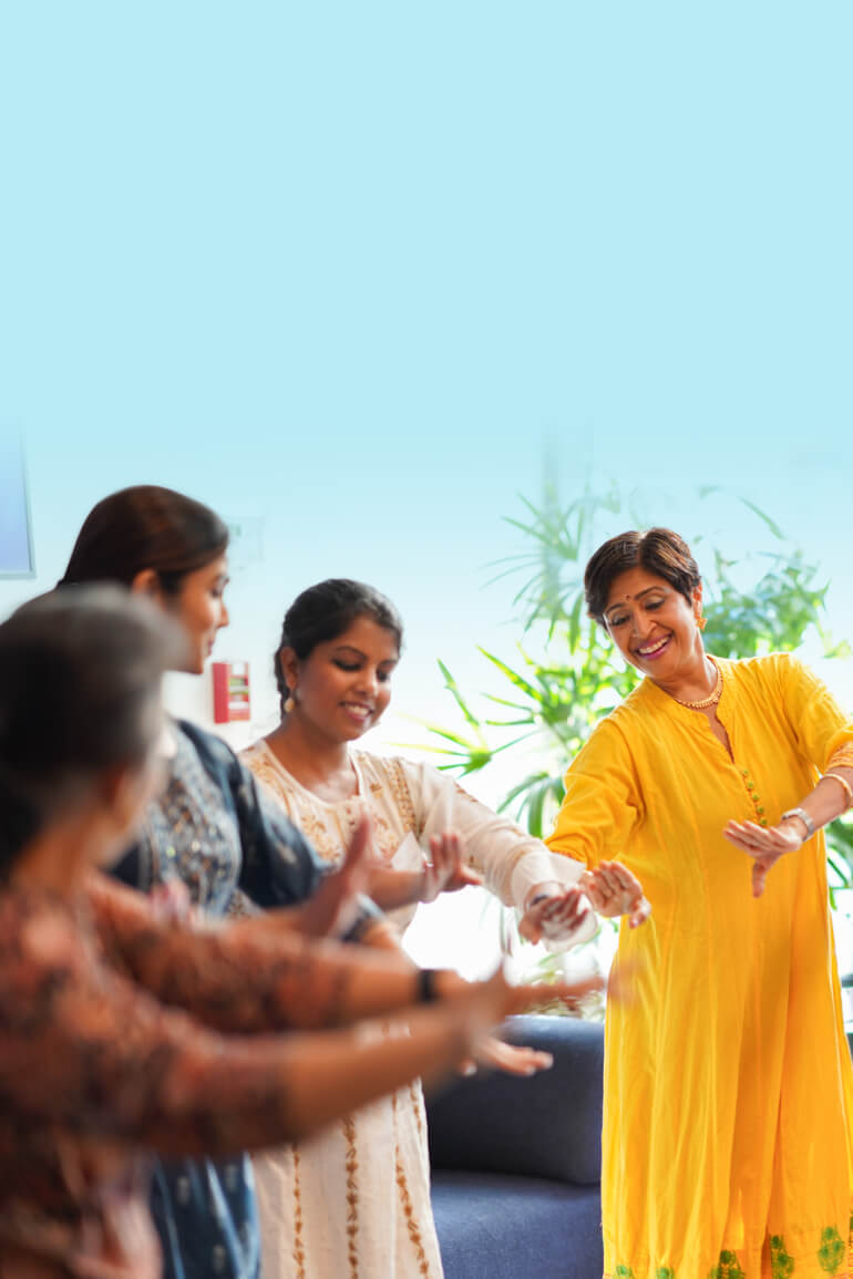 A group of women in traditional attire dance together, with one woman in a yellow dress leading and smiling, set against a light blue background.