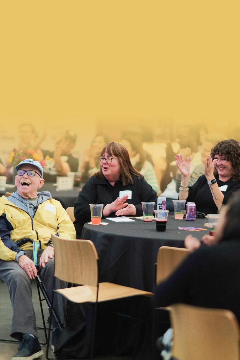 A man in a yellow jacket and gray baseball cap looks up and smiles as the people around him cheer and clap.